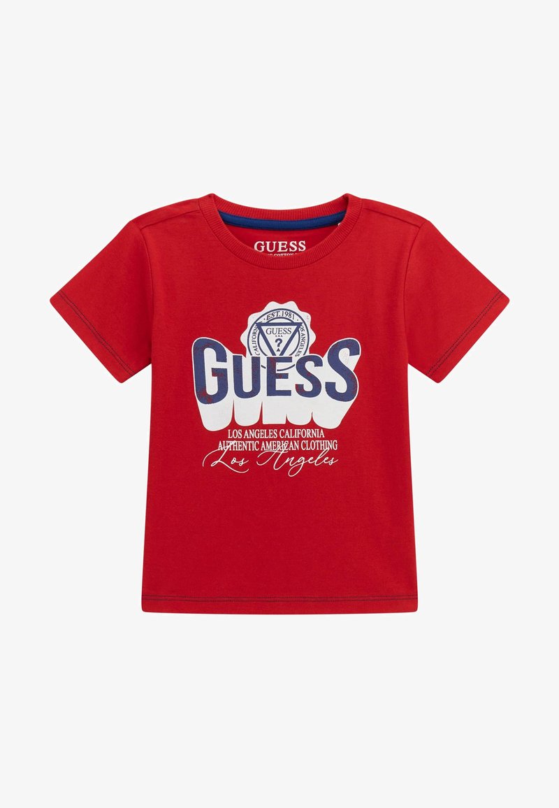 Rotes Baumwoll-T-Shirt mit kurzen Ärmeln, das mit großen weißen und blauen "GUESS"-Schriftzügen, einem runden Logo und zusätzlichen weißen Textdetails versehen ist.