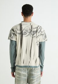 Chemise beige clair à manches courtes avec un motif abstrait gris, superposée à un haut à manches longues bleu-vert. Portée avec un jean à motifs de couleur claire.