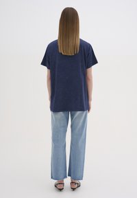 Tamsiai mėlynas per didelis T-shirts su trumpomis rankovėmis, derinamas su šviesiai mėlynomis plačiomis džinsų kelnėmis su nusidėvėjusiomis briaunomis, dėvimas su juodais sandalais.