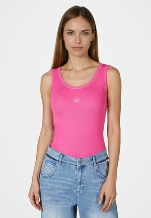 Woman with straight brown hair wearing a pink sleeveless top and light blue jeans, standing against a white background.