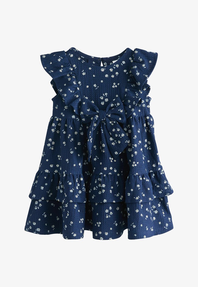 Robe bleu marine sans manches avec imprimé de marguerites blanches, épaules volantées, jupe à étages et grand nœud noué à la taille.