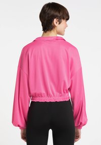 Afskåret, langærmet pink jakke med et struktureret, åndbart stof. Har krave og elastisk kant. Båret over sorte tætsiddende bukser.