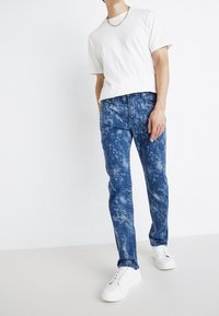 Lehké bílé bavlněné tričko v kombinaci s vybledlými modrými džíny s vzorem připomínajícím mraky, doplněné o bílé tenisky.