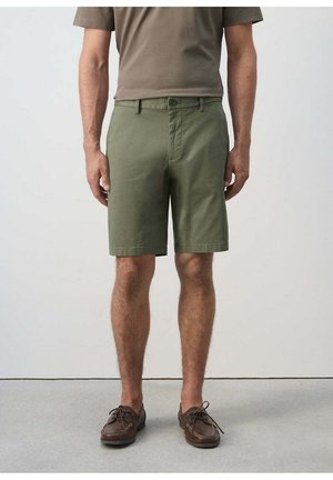 Hombre con pantalones cortos verde oliva, camisa marrón y zapatos de piel marrón con cordones, de pie sobre un suelo gris frente a una pared lisa y clara.