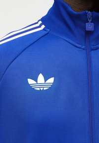 Blå sportjakke med lynlås, hvid Adidas trefoil-logo på brystet og tre hvide striber på skulderen.