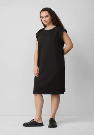 Schwarzes Kleid mit kurzen, umgeschlagenen Ärmeln und geradem Schnitt. Hergestellt aus glattem Stoff mit einfachem Ausschnitt. Getragen mit schwarzen Slippern.