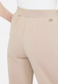 Pantaloni bej cu talie elastică, având o textură netedă, cu un detaliu subtil din metal auriu. Croială ajustată, cu un design lejer pe picior.