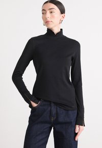 Zwarte lange mouwen turtleneck top met een pasvorm ontwerp, een gladde textuur en zijritsen bij de manchetten, gecombineerd met donkerdenim broek.