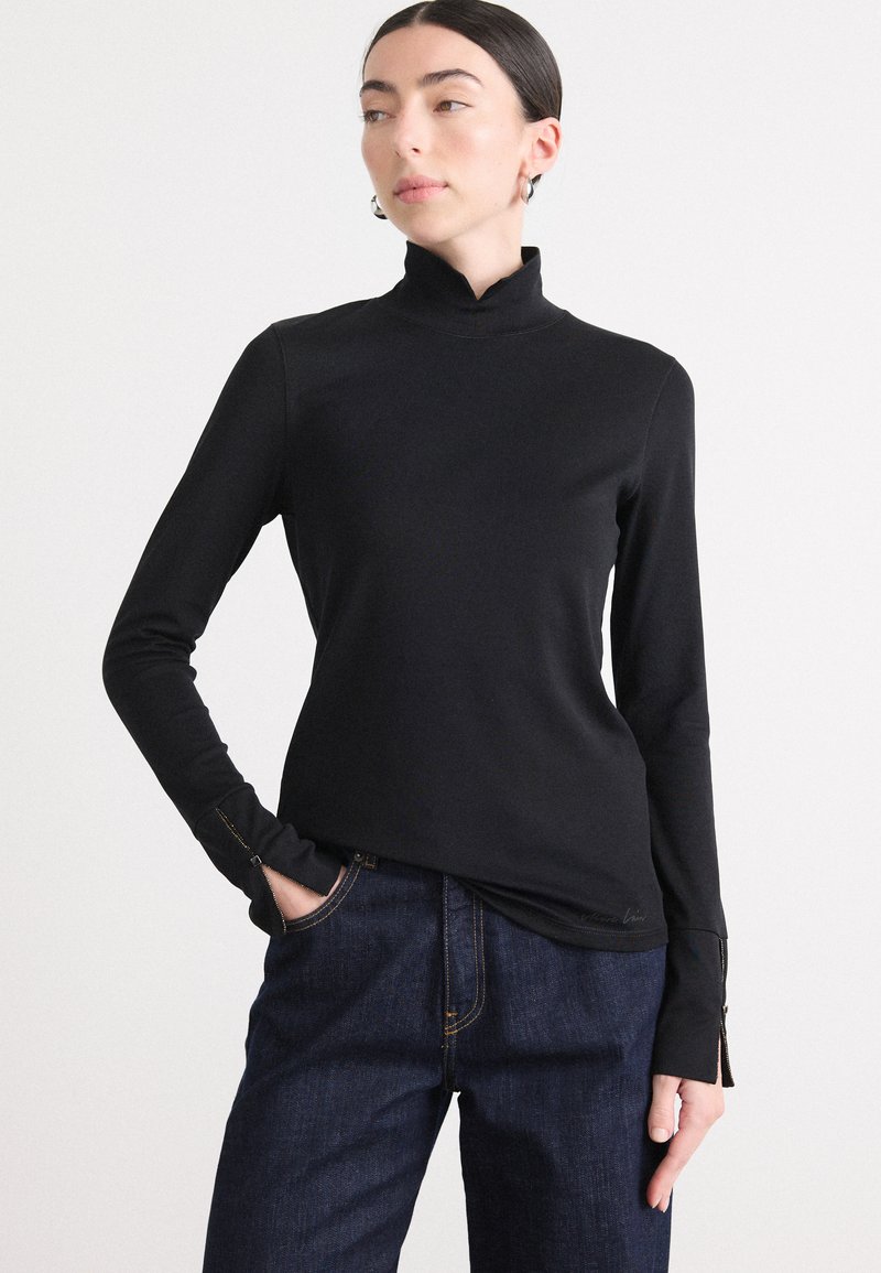 Zwarte lange mouwen turtleneck top met een pasvorm ontwerp, een gladde textuur en zijritsen bij de manchetten, gecombineerd met donkerdenim broek.