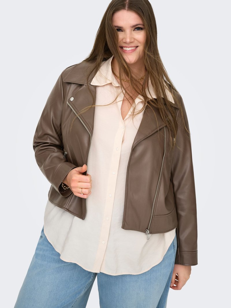 L ächelnde Frau mit langen Haaren, die eine braune Lederjacke über einem cremefarbenen Hemd und hellblauen Jeans trägt, vor einem einfarbigen Hintergrund.