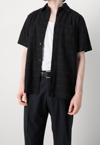 Zwarte korte mouwen shirt met een gestructureerd patroon, knopen aan de voorkant en een borstzak, gecombineerd met zwarte broek en een riem.