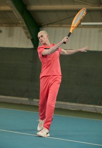 Tennis spiller i lys koral sportsbeklædning, der udfører et sving med en ketsjer. Har en matchende top og bukser, med hvide sneakers og en grøn baggrund.