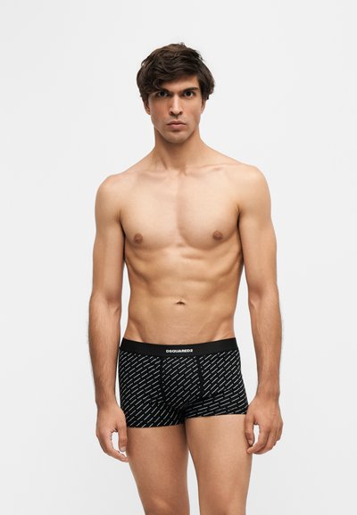 Homme torse nu debout devant un fond uni portant un boxer noir à motifs avec une ceinture "DSQUARED2".