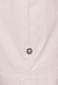 Jemný růžový materiál v detailu, na němž je kulatý kovový knoflík s květinovým vzorem a prošíváním podél švu.