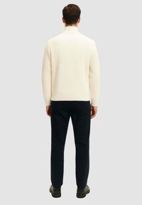 Maglione a collo alto a coste color crema con una silhouette aderente, abbinato a pantaloni blu. Il tessuto sembra morbido con un motivo testurizzato.