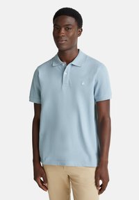 Mladý muž oblečený v světle modré polo tričko s krátkým rukávem a béžových kalhotách, stojící před jednoduchým světlým pozadím.