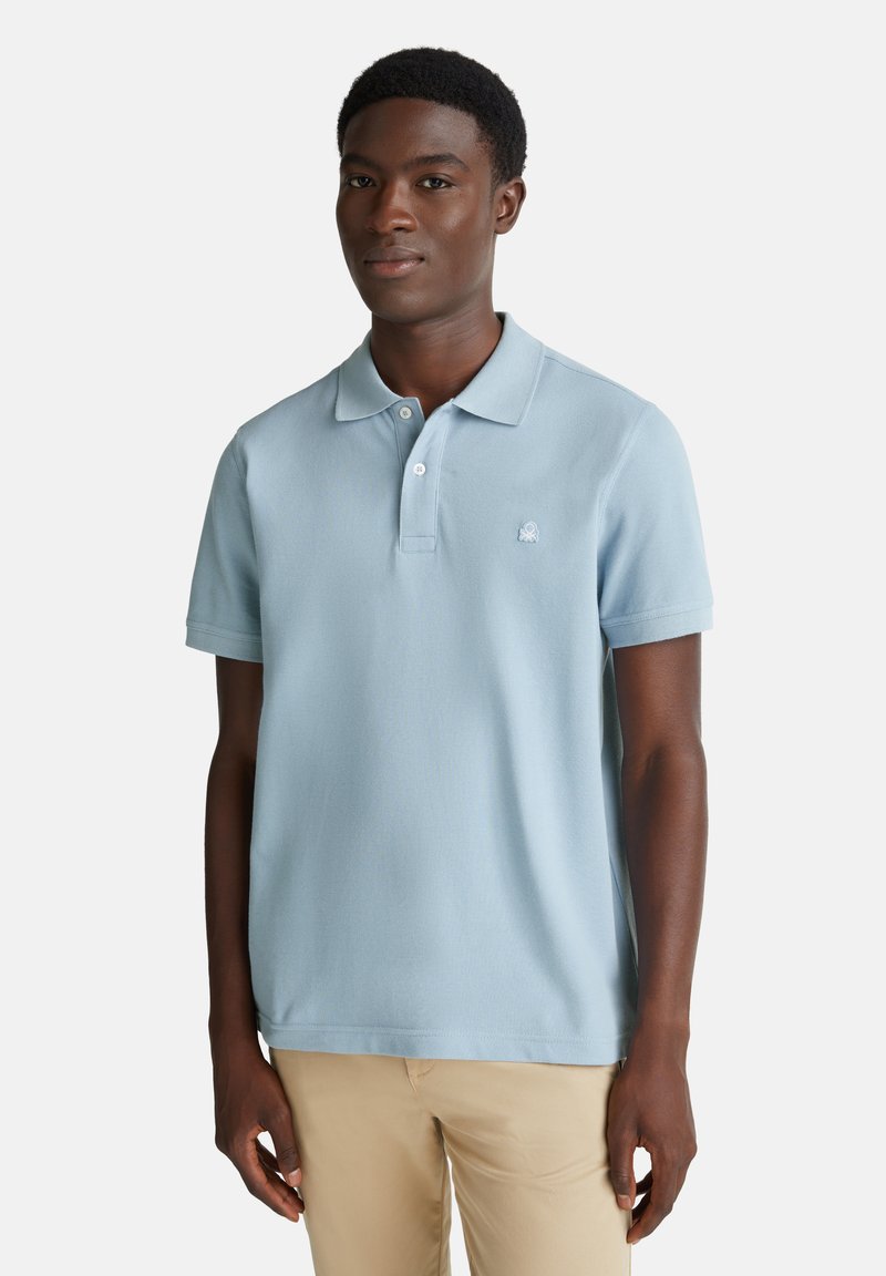 Mladý muž oblečený v světle modré polo tričko s krátkým rukávem a béžových kalhotách, stojící před jednoduchým světlým pozadím.