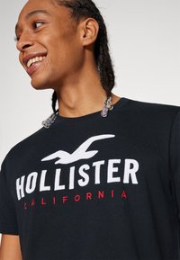 Melns kokvilnas t-krekls ar baltu "HOLLISTER" tekstu un logo ar sarkaniem akcentiem. Arapa aplis un gluda tekstūra, piemērots ikdienas valkāšanai.