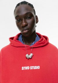 Jong persoon met gevlochten haar, die een rode STWD STUDIO hoodie, een blauw gestreept shirt en een zilveren ketting draagt tegen een plain witte achtergrond.