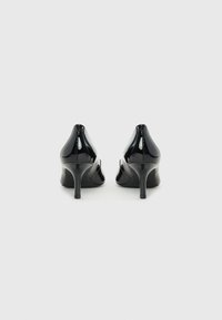 Tacones altos de charol negro con punta afilada, tacón de aguja delgado y textura suave. Diseño de atrás abierto con detalles de costura mínimos.