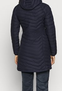 Marinblå vadderad jacka med chevron-sömmar, en huva och en figursydd midja. Baksidan har vertikala sömmar för extra struktur.