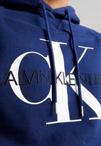 Felpa blu/navy con un grande logo bianco raffigurante "CK" e "CALVIN KLEIN JEANS" ricamato in nero. I lacci del cappuccio hanno punte in metallo.