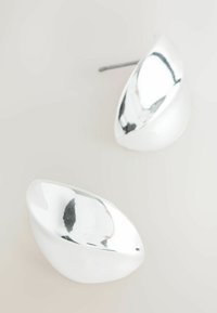 Cercei din metal argintiu cu un design neted, oval și ușor curbat. Suprafața este strălucitoare, cu un finisaj reflectant.