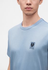 Νέος άνδρας που φοράει ένα ανοιχτό μπλε t-shirt Belstaff με ένα μικρό λογότυπο στο στήθος, στέκεται μπροστά σε ένα απλό λευκό φόντο.
