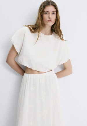 Mujer joven con cabello largo y ondulado, vistiendo un top corto blanco y suelto y una falda de cintura alta con sutiles patrones florales, de pie frente a una pared blanca.