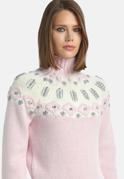 Młoda kobieta z brązowymi włosami w krótkim bobie, ubrana w różowo-biały sweter z golfem, ozdobiony srebrnymi koralikami i kamieniami szlachetnymi.