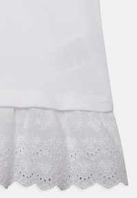 Camicia bianca in cotone con orlo decorativo in pizzo ad occhiello. Presenta una parte superiore texturizzata con una finitura liscia e bordi ondulati.