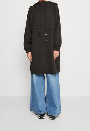 Personne portant un long manteau noir à capuche par-dessus un jean bleu à jambes larges et des chaussures de sport noires avec des détails verts et blancs.