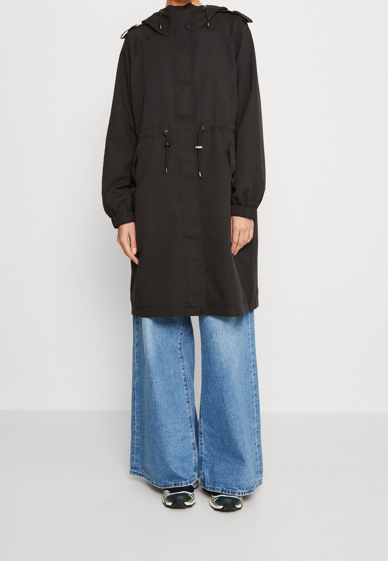 Personne portant un long manteau noir à capuche par-dessus un jean bleu à jambes larges et des chaussures de sport noires avec des détails verts et blancs.