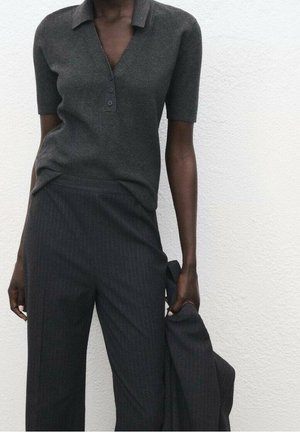 Personne portant un haut gris foncé à manches courtes avec col et boutons et un pantalon large à fines rayures, tenant une veste assortie dans une main.