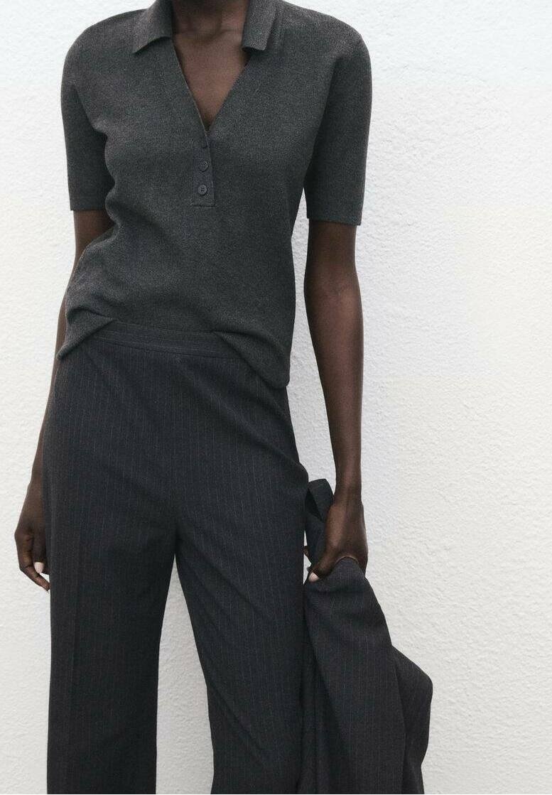 Personne portant un haut gris foncé à manches courtes avec col et boutons et un pantalon large à fines rayures, tenant une veste assortie dans une main.