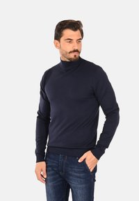 Maglione dolcevita blu navy, polsini e orlo a coste, texture a maglia liscia, indossato con jeans di denim scuro, in piedi con le mani nelle tasche.