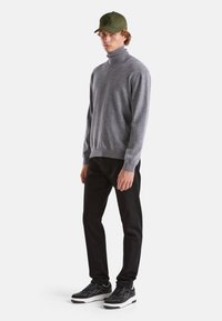 Maglione grigio a collo alto realizzato in materiale morbido, pantaloni neri e sneakers nere con suola bianca e accenti grigi. Un berretto verde oliva completa il look.