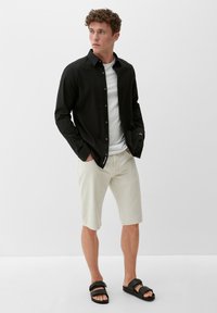 Schwarze Hemdbluse, weißes T-Shirt darunter, beiges Shorts und schwarze Sandalen; legere Kleidung mit einfarbigen Tönen und schlichtem Design.