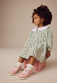 Robe en vichy vert avec fleurs brodées, col blanc et boutons. Chaussures Mary Jane en cuir rose avec boucle et chaussettes blanches bordées de dentelle.
