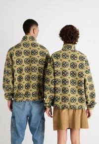 Jachete din fleece galbene cu modele florale marine rotunde. Guler ridicat și manșete elastice. Una este asortată cu o fustă de catifea ciocănită maro.