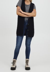 Schwarzer, gestrickter langer Weste mit Knöpfen, kombiniert mit einem grauen T-Shirt, blauen Skinny-Jeans und beigen Schnürstiefeln mit schwarzen Sohlen.