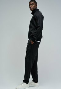 Chaqueta deportiva negra con cuello alto y cremalleras, combinada con pantalones negros que tienen bolsillos laterales, complementados con zapatillas blancas.