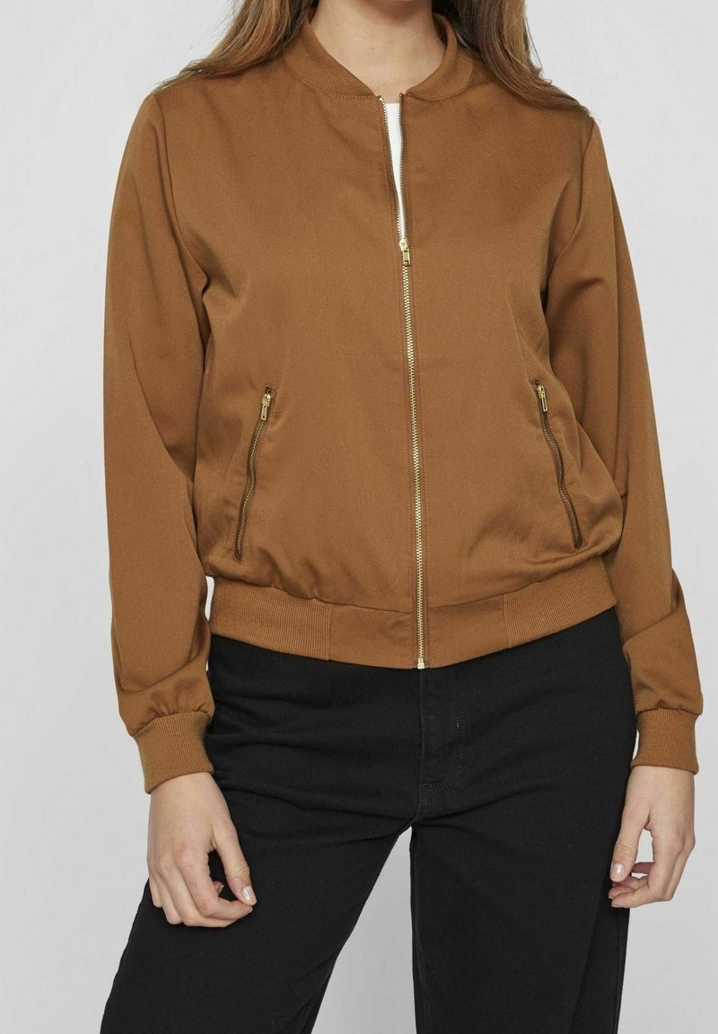 Femme portant une veste marron à fermeture éclair avec poignets et ourlet côtelés, associée à un pantalon noir, posant devant un fond uni.