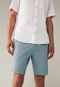 Helleblaue Shorts aus glattem Stoff, mit Seitentaschen und geradem Schnitt. Kombiniert mit einem weißen, kurzärmligen Leinenhemd mit Knöpfen.