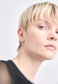 Srebrna naušnica sa prozirnim kristalom. Crni top s perlicama. Krupni plan uha i vrata, ističući teksture i detalje.