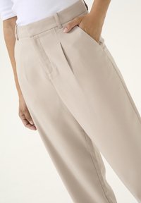Pantalon beige ajusté avec plis, taille haute, poches latérales et texture lisse. Main posée sur la poche, mettant en valeur les détails du vêtement.