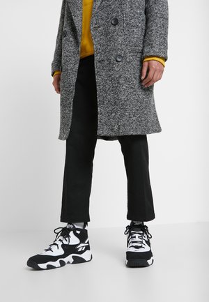 Muž s černo-bílými vysokými teniskami, zkrácenými černými kalhotami, hořčicovým svetrem a šedým strukturovaným kabátem stojící na bílé podlaze.