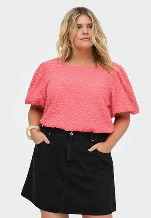 Femeie purtând o bluză roz texturată cu mâneci bufante, băgată într-o fustă neagră de denim, cu mâinile în buzunare, stând în fața unui fundal simplu.