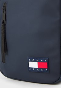 Navy blue bag with a textured surface and a black zip. Features a logo patch with red, white, and blue colors, marked "TOMMY JEANS."