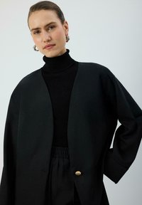 Melna pārmērīgi liela jaka ar V-veida kakla izgriezumu, vienu zelta pogu un platiem piedurknēm, valkāta pār melnu augstu apkakli. Gluda tekstūra.